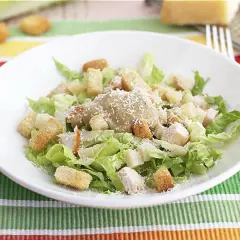 Una deliciosa ensalada cesar con pollo, crutones crujientes y queso parmesano rallado, servida en un plato blanco sobre un mantel colorido.