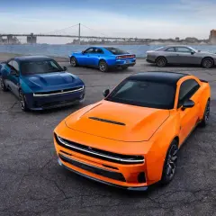 Pomarańczowy i niebieski Dodge Charger Hellcat oraz szary coupe stoją nad wodą z mostem w tle.