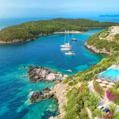 Vacker vy över Sivota centrum med turkost vatten, segelbåtar och ett lyxigt hotell med poolområde.