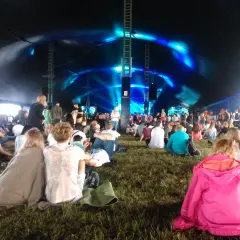Widownia siedzi na trawie pod namiotem, czekając na koncert. Niebieskie światła oświetlają scenę, tworząc atmosferę festiwalu Open'er.