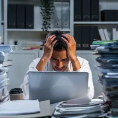 Mężczyzna z rękami na głowie siedzi przy biurku zasypanym papierami, czując ogromną frustrację.