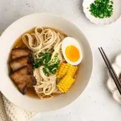 A miseczce shoyu ramen z makaronem, plastrami boczku, jajkiem na pół, kukurydzą i szczypiorkiem. Obok pałeczki i dodatkowy boczek.