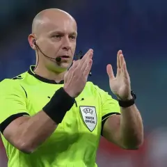Sędzia piłkarski w żółtej koszulce z emblematem UEFA, z mikrofonem przy uchu, pokazuje gest rękami. Ciekawe, ile zarabia sędzia piłkarski za takie emocje.