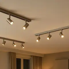 Dos rieles con focos de techo modernos, de metal cepillado, iluminan un espacio acogedor con cortinas y una ventana.