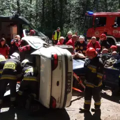 Akcja ratunkowa ksrg po wypadku. Strażacy wyciągają poszkodowanych z przewróconego samochodu. W tle wozy strażackie i karetka.