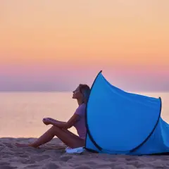 Kobieta siedzi na plaży przy rozłożonym namiocie plażowym. Zachód słońca nad morzem. Jak złożyć namiot plażowy instrukcja.