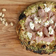 Pizza mit Mortadella, Pistazien und cremigem Käse, daneben einige Pistazien und eine Zitronenspalte.