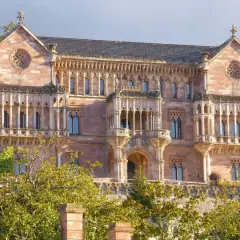 El **Palacio de Comillas**, una joya arquitectónica de piedra rosada, se alza majestuoso entre la vegetación, con sus balcones y rosetones detallados.
