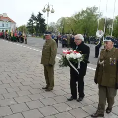Trzech mężczyzn w mundurach wojskowych i cywilnych, jeden z nich niesie wieniec. W tle parada i orkiestra.