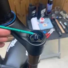 A hand uses a green hex wrench to adjust a bolt on a black bicycle fork, part of a Fox 34 service. Tools and lubricants are visible in the background.