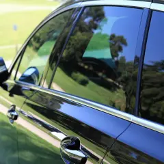Czarne auto z przyciemnianiem przednich szyb, odbijające zieleń trawy i drzew.