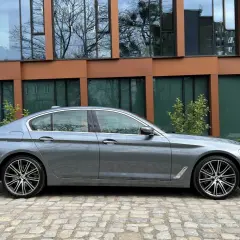 Szary sedan BMW M5 G30 stoi na brukowanym podjeździe przed nowoczesnym budynkiem z brązowymi ramami okien.