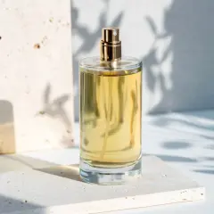 Szklana butelka perfum z złotą nakrętką na kamiennej podstawie, w cieniu liści.