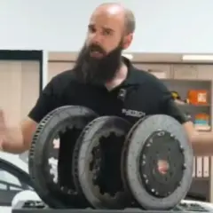 Hombre con barba muestra tres discos de freno, uno de ellos con perforaciones, para mejorar la refrigeración en un coche deportivo.
