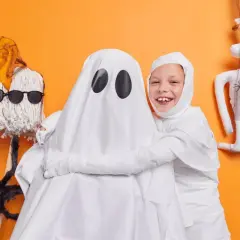 Dzieci w strojach mumii i ducha, świetny pomysł, jak zrobić strój na Halloween. Obok pluszowy szkielet i pająk.