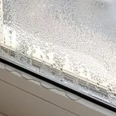 Gotas de agua cubren el cristal de la ventana, indicando condensación. El alféizar muestra pequeñas acumulaciones de agua.