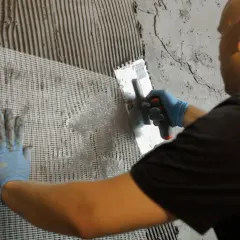 Hand mit blauer Handschuh drückt Gittergewebe in Mörtel auf der Wand. Dies hilft gegen Feuchtigkeit in der Wand.