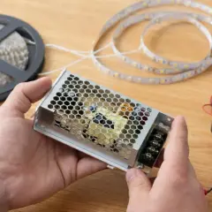 Ręce trzymają zasilacz do taśmy LED. W tle szpulka z taśmą i miernik. Nowoczesna technologia LED gotowa do montażu.