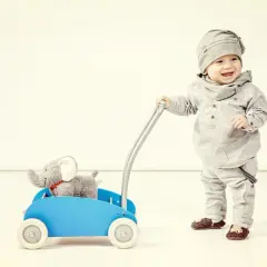 Uśmiechnięte dziecko pcha niebieski wózek z pluszowym słonikiem. To świetny pchacz dla dziecka, wspierający fizjoterapię i pierwsze kroki.