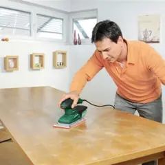 Un homme utilise une ponceuse vibrante ou excentrique pour rénover une table en bois.