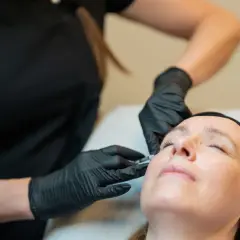 En behandlare med svarta handskar ger en kvinna fillers innehåll i ansiktet.