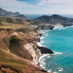 Costa escarpada cerca de Níjar, con el mar turquesa rompiendo en acantilados blancos y rojizos. Montañas verdes y una carretera serpentean en la distancia.