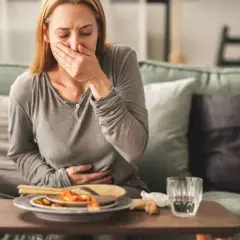 Kobieta siedzi na kanapie, trzymając się za brzuch i zakrywając usta, cierpiąc z powodu niestrawności. Na stoliku resztki jedzenia i szklanka wody.