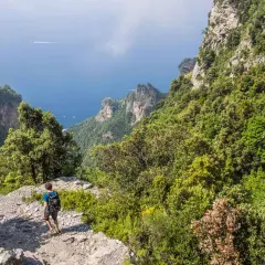 A hiker walks along the rocky path of the Sentiero degli Dei, with lush green mountains and the blue sea stretching out below.
