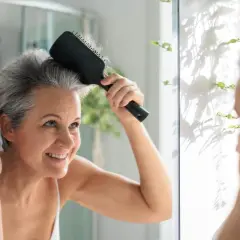 Mujer sonriente se peina el cabello gris en el espejo, lista para matizar canas y lucir radiante.