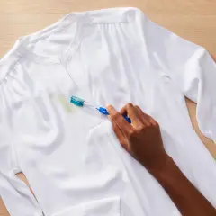 Une main utilise une brosse à dents pour enlever une tache verte sur une chemise blanche. Astuce : comment enlever de la colle sur un vêtement.