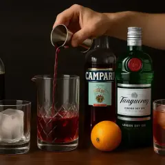 Dłoń wlewa czerwony płyn z jiggera do szklanicy. Obok butelki ginu, Campari, pomarańcza i gotowy koktajl.