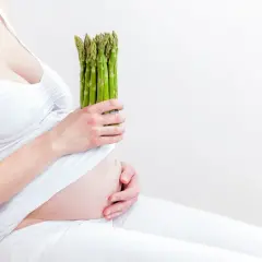 Kobieta w ciąży trzyma pęczek zielonych szparagów, symbol zdrowej diety.