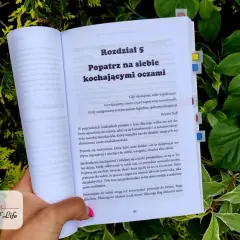 Otwarta książka "Popatrz na siebie kochającymi oczami" w otoczeniu zieleni. Tekst mówi o walce z negatywną samokrytyką i brakiem akceptacji siebie.