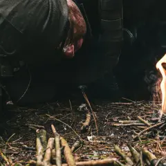 Osoba w rękawicach rozpala ognisko w lesie. Dym unosi się nad płomieniami, a wokół leżą gałązki. To cenne sere b porady dla każdego survivalowca.