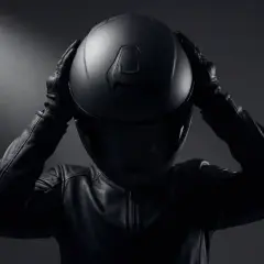 Persona con chaqueta de cuero y guantes se pone un casco negro, con líneas afiladas y modernas.