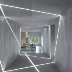 Nowoczesny korytarz z betonowymi ścianami i geometrycznym podświetleniem ściany od góry. W tle salon z kominkiem i kanapą.