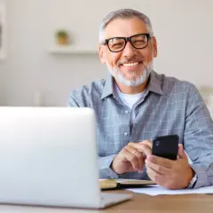 Uśmiechnięty senior z okularami pracuje przy laptopie i telefonie. To ulga dla pracujących seniorów, którzy cenią sobie nowoczesne rozwiązania.