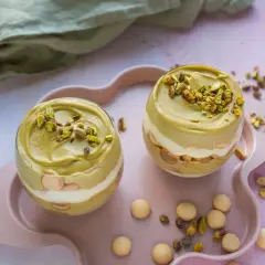 Dwa pucharki z deserem: krem pistacjowy z mascarpone, przełożony biszkoptami i bitą śmietaną, posypany pistacjami.