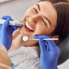 Uśmiechnięta pacjentka podczas zabiegu sanacji jamy ustnej. Dentysta w niebieskich rękawiczkach używa wiertła i lusterka.