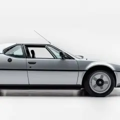Srebrne BMW M1 E26 widziane z boku, z otwartymi drzwiami.