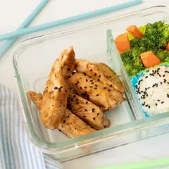 A delicious chicken bento box with teriyaki chicken strips, white rice sprinkled with black sesame seeds, and steamed broccoli with carrots.