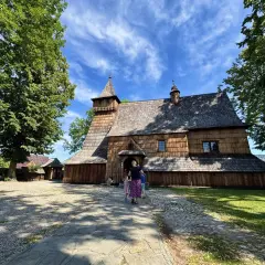 Drewniany kościół w Dębnie, z charakterystyczną wieżą i gontowym dachem, otoczony zielenią.