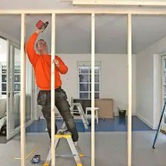 Man i orange tröja monterar träreglar för en ny vägg med skruvdragare.