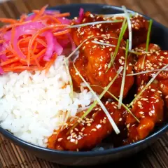 En skål med ris, krispigt koreanskt friterad kyckling i en söt och kryddig sås, toppad med sesamfrön och salladslök. Serveras med en sida av picklad morot och rödlök.