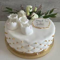 Delikatny tort na chrzest dla Julii, ozdobiony białymi bucikami, kwiatami i złotymi perełkami.