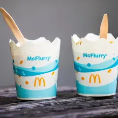 Dwa kubeczki McFlurry z McDonald's, każdy z drewnianą łyżeczką, stoją na drewnianym blacie.