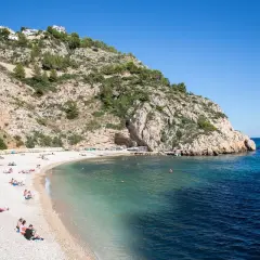 Gente disfrutando del sol en una playa de guijarros en la **Costa Blanca**, con aguas turquesas y un acantilado rocoso.