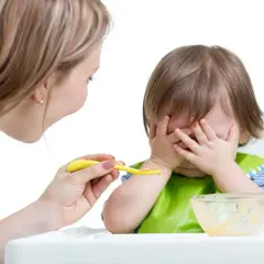 Mama próbuje nakarmić malucha, ale niemowlę nie chce jeść, zakrywając buzię rączkami.