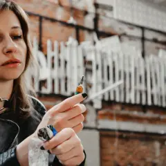 Kobieta z kolczykiem w kształcie serca na szyi trzyma strzykawkę. Jej zęby, choć nie widać ich wyraźnie, mogą być symbolem zniszczeń, jakie niesie ze sobą życie narkomana.