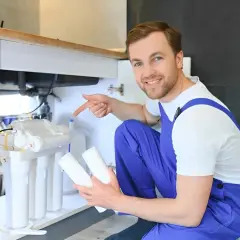 Un technicien souriant montre les cartouches pour une installation filtre à eau maison sous un évier.